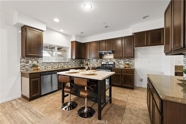 kitchen featuring stone counters, under cabinet range hood, stainless steel appliances, dark brown cabinets, and backsplash