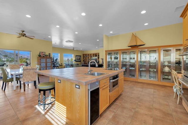 kitchen with stainless steel oven, ceiling fan, sink, tile countertops, and an island with sink