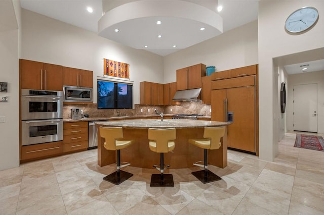 kitchen featuring a high ceiling, tasteful backsplash, built in appliances, an island with sink, and a breakfast bar area