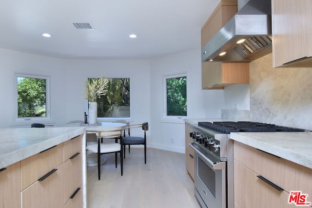 kitchen featuring high end stainless steel range, wall chimney exhaust hood, plenty of natural light, and light brown cabinets