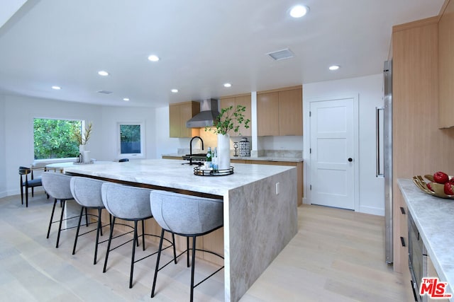 kitchen featuring wall chimney exhaust hood, a breakfast bar, light brown cabinets, a center island with sink, and light hardwood / wood-style floors