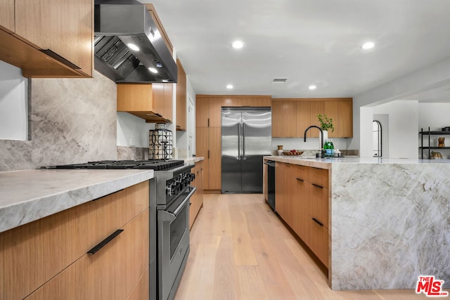 kitchen featuring light stone countertops, wall chimney exhaust hood, premium appliances, tasteful backsplash, and light hardwood / wood-style flooring