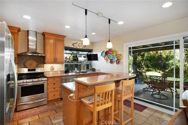 kitchen with light stone countertops, appliances with stainless steel finishes, wall chimney exhaust hood, a breakfast bar, and hanging light fixtures