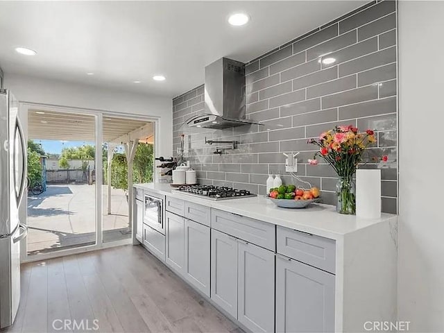 kitchen with light wood-type flooring, stainless steel appliances, backsplash, and wall chimney range hood