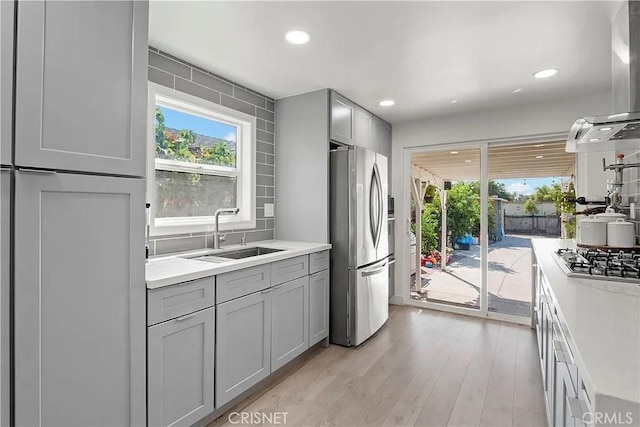 kitchen featuring appliances with stainless steel finishes, gray cabinetry, wall chimney range hood, sink, and light hardwood / wood-style flooring