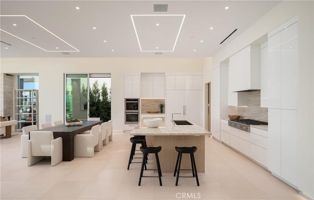 kitchen featuring light stone countertops, a large island with sink, tasteful backsplash, and white cabinetry