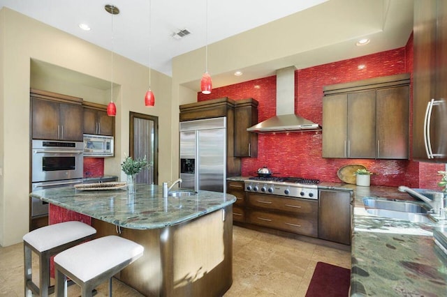 kitchen featuring wall chimney range hood, decorative backsplash, built in appliances, dark stone counters, and a kitchen island with sink