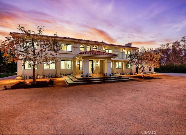 back of house at dusk with a balcony, a tile roof, and stucco siding