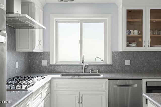 kitchen featuring glass insert cabinets, white cabinetry, a sink, wall chimney exhaust hood, and stainless steel appliances