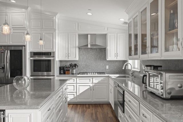 kitchen featuring appliances with stainless steel finishes, white cabinets, decorative light fixtures, wall chimney exhaust hood, and glass insert cabinets