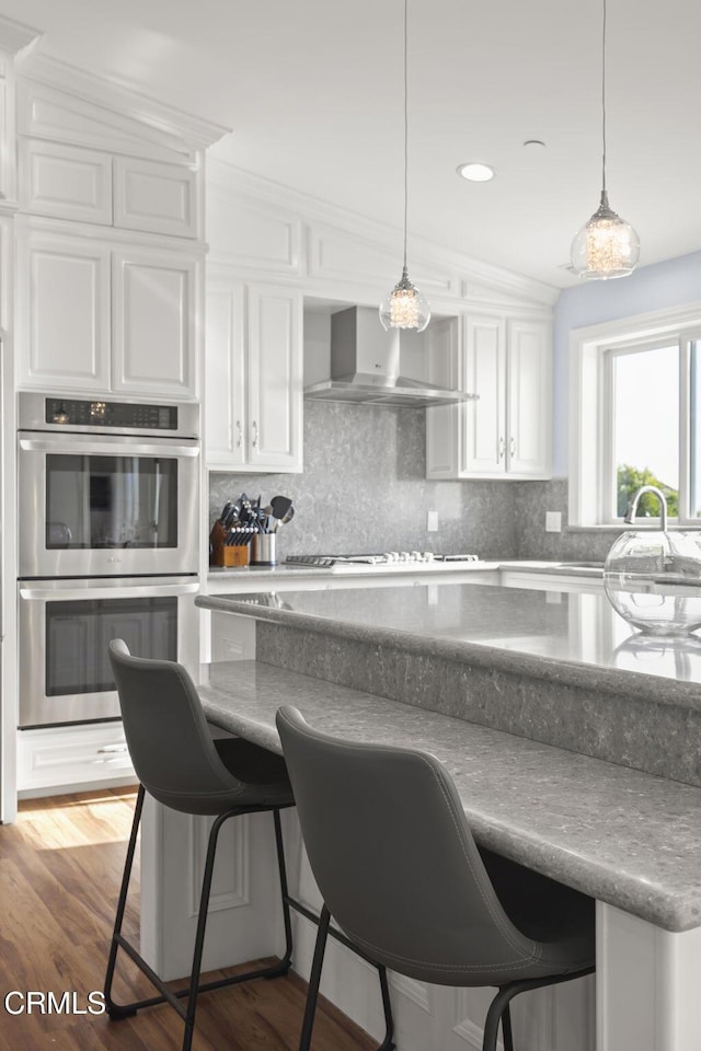 kitchen with white cabinetry, a breakfast bar, wall chimney range hood, and stainless steel double oven