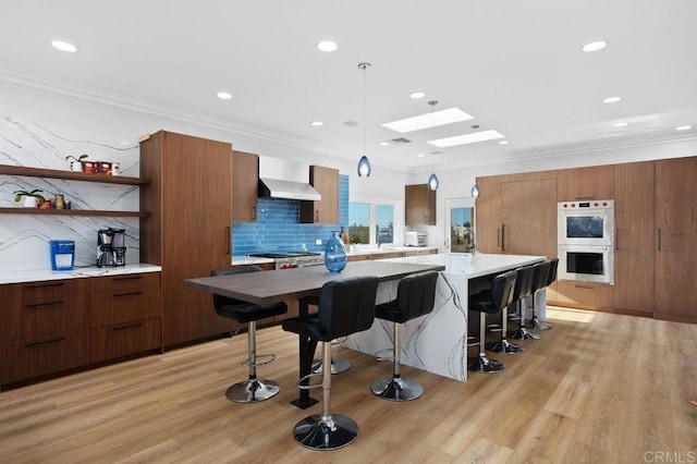 kitchen with hanging light fixtures, wall chimney range hood, double oven, light hardwood / wood-style floors, and decorative backsplash