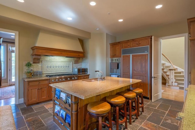 kitchen with decorative backsplash, appliances with stainless steel finishes, premium range hood, sink, and an island with sink