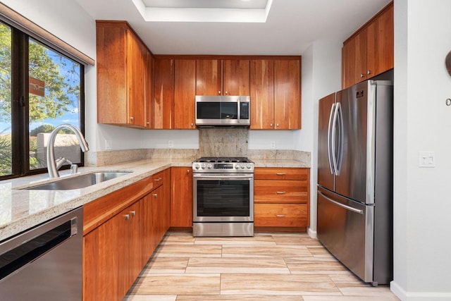 kitchen with light stone countertops, stainless steel appliances, a tray ceiling, sink, and light hardwood / wood-style floors