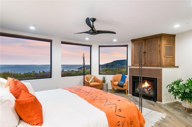 bedroom with a tile fireplace, a water view, ceiling fan, light wood-type flooring, and multiple windows