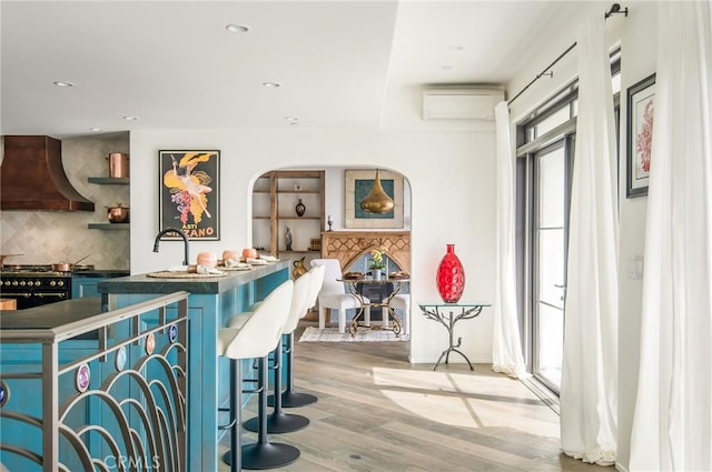 kitchen with light wood-type flooring, backsplash, custom range hood, a wall unit AC, and stainless steel range with gas cooktop