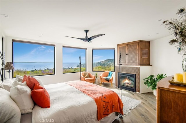 bedroom with a water view, ceiling fan, light hardwood / wood-style floors, and a tiled fireplace