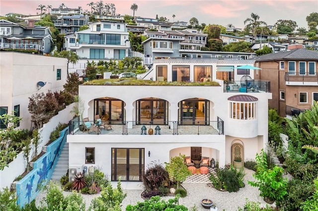 back of property at dusk with a balcony, a residential view, stucco siding, and french doors