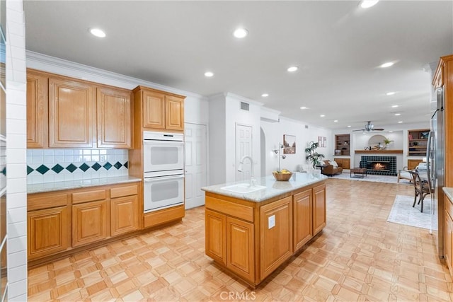 kitchen featuring backsplash, ceiling fan, double oven, sink, and an island with sink