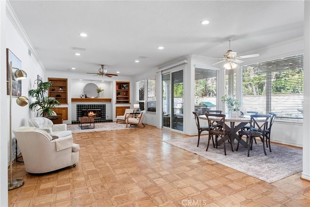 living room with crown molding, ceiling fan, built in shelves, a fireplace, and light parquet flooring