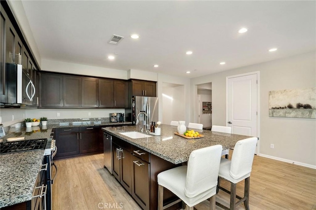 kitchen with light stone counters, stainless steel appliances, a kitchen island with sink, sink, and light hardwood / wood-style floors