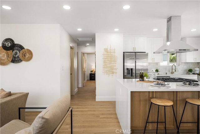 kitchen with appliances with stainless steel finishes, white cabinetry, backsplash, island exhaust hood, and light wood-type flooring