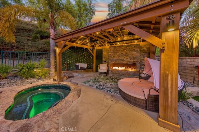 pool at dusk featuring an in ground hot tub, a patio area, an outdoor stone fireplace, and a gazebo