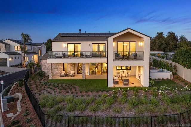 back house at dusk with solar panels, a balcony, outdoor lounge area, a yard, and a patio