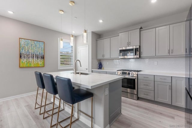 kitchen with gray cabinets, a center island with sink, decorative backsplash, hanging light fixtures, and appliances with stainless steel finishes