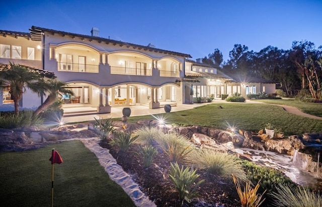 back house at dusk with a lawn, a balcony, and a patio