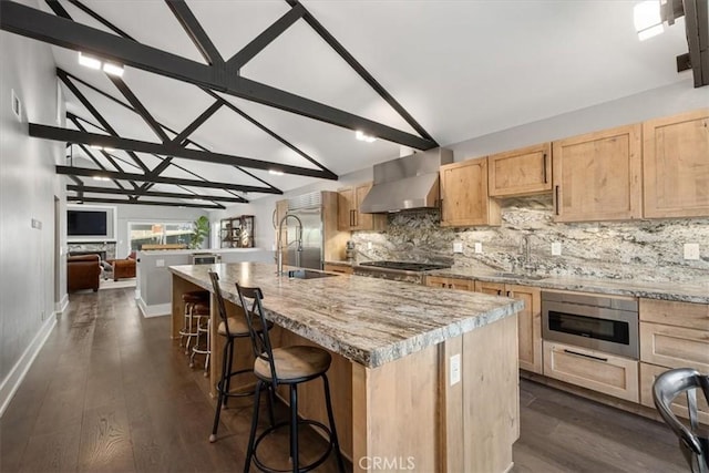 kitchen featuring wall chimney exhaust hood, a kitchen bar, light brown cabinetry, vaulted ceiling with beams, and an island with sink