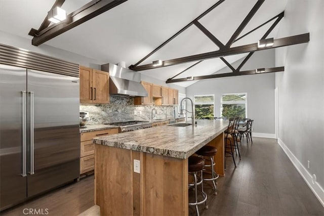 kitchen with wall chimney range hood, sink, a kitchen island with sink, vaulted ceiling with beams, and stainless steel appliances