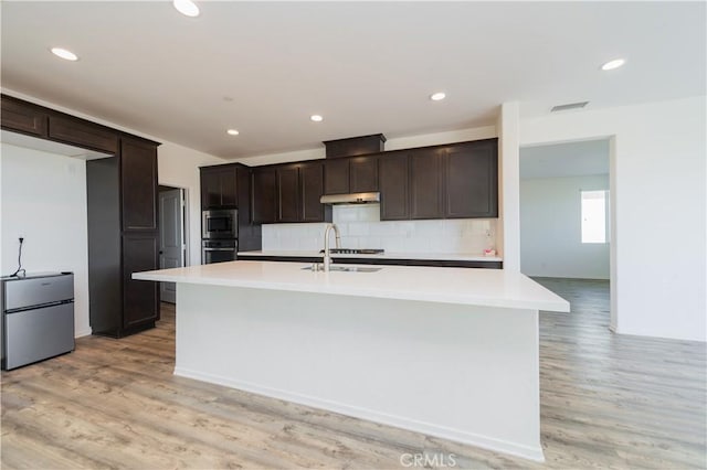 kitchen with decorative backsplash, light wood-type flooring, stainless steel appliances, sink, and a center island with sink