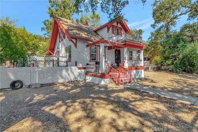 view of front facade featuring a balcony, covered porch, a shingled roof, fence, and a chimney