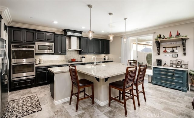 kitchen with wall chimney exhaust hood, a kitchen bar, appliances with stainless steel finishes, an island with sink, and pendant lighting