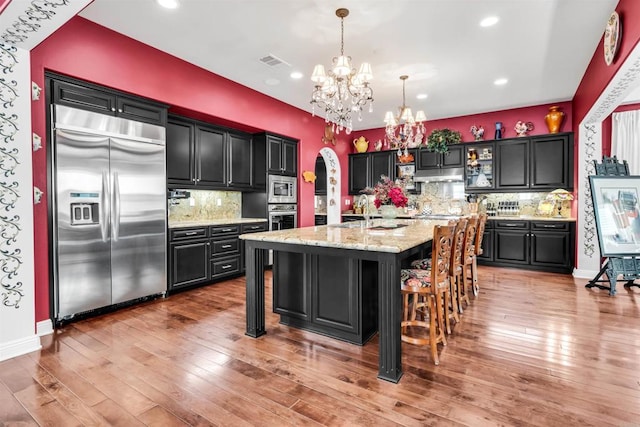 kitchen featuring pendant lighting, hardwood / wood-style floors, backsplash, built in appliances, and an island with sink