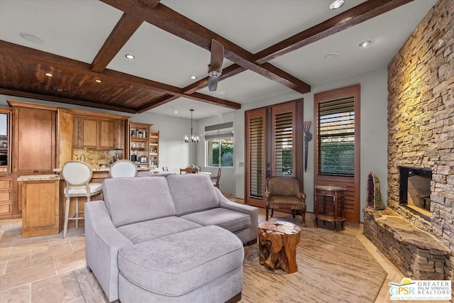 living room with ceiling fan with notable chandelier, beam ceiling, a stone fireplace, and coffered ceiling