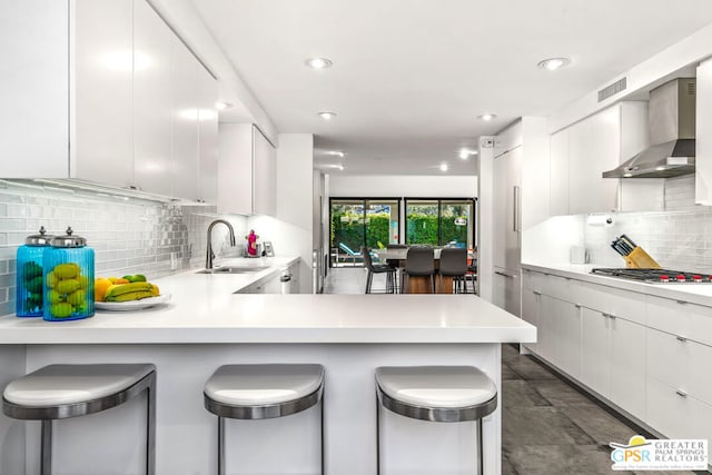 kitchen featuring kitchen peninsula, a kitchen breakfast bar, wall chimney exhaust hood, sink, and white cabinets