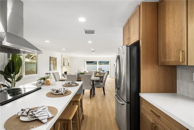 kitchen featuring brown cabinets, light countertops, visible vents, freestanding refrigerator, and wall chimney range hood