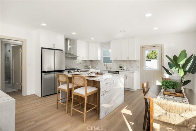 kitchen featuring wall chimney range hood, stainless steel fridge, a breakfast bar area, a center island, and white cabinets