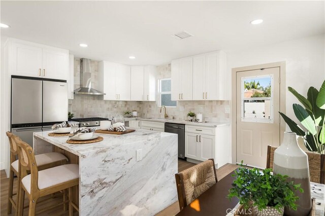 kitchen featuring appliances with stainless steel finishes, light stone counters, white cabinets, a kitchen island, and wall chimney exhaust hood