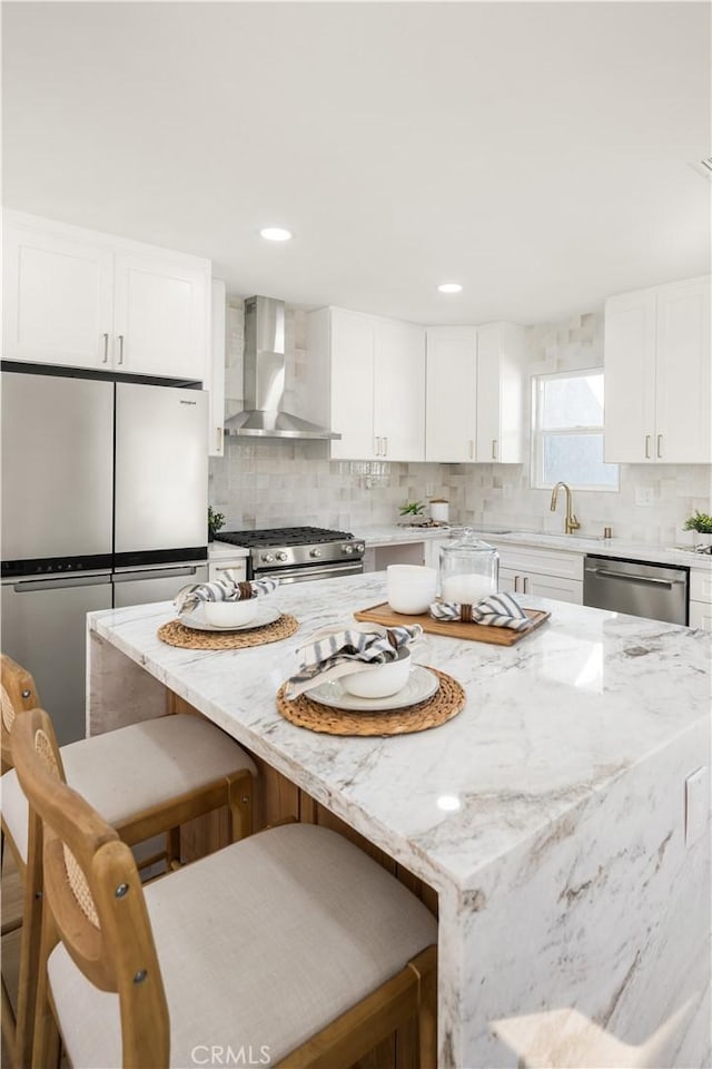 kitchen with wall chimney range hood, appliances with stainless steel finishes, white cabinetry, a kitchen breakfast bar, and light stone countertops