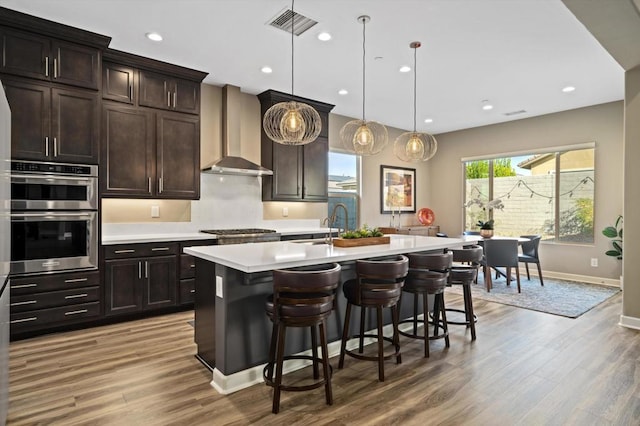 kitchen with pendant lighting, a breakfast bar, dark brown cabinetry, a center island with sink, and wall chimney exhaust hood