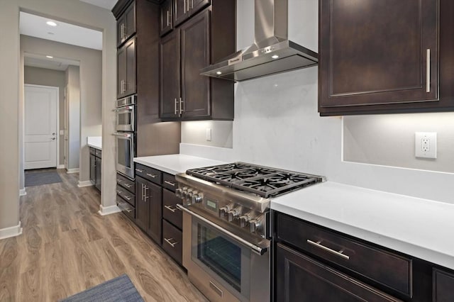 kitchen with dark brown cabinetry, stainless steel appliances, light hardwood / wood-style flooring, and wall chimney range hood