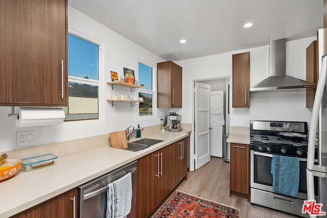 kitchen featuring sink, wall chimney exhaust hood, appliances with stainless steel finishes, stacked washer / drying machine, and light hardwood / wood-style floors