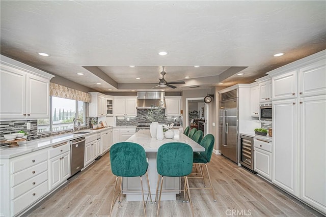 kitchen featuring built in appliances, a tray ceiling, white cabinets, beverage cooler, and wall chimney exhaust hood