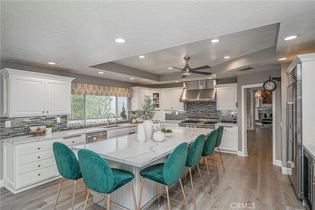 kitchen with a tray ceiling, light stone countertops, white cabinets, a kitchen island, and wall chimney exhaust hood