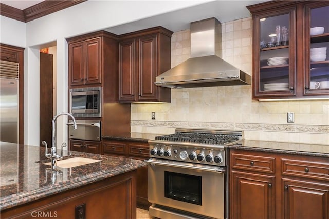 kitchen with wall chimney exhaust hood, dark stone counters, built in appliances, tasteful backsplash, and sink