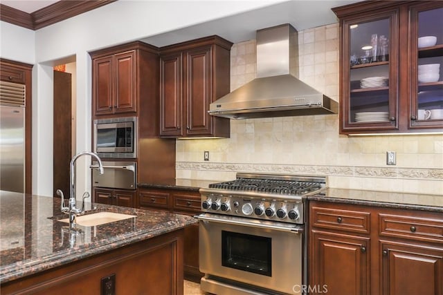 kitchen featuring backsplash, glass insert cabinets, a sink, built in appliances, and wall chimney exhaust hood