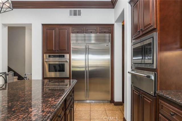 kitchen with light tile patterned floors, dark stone counters, built in appliances, and ornamental molding